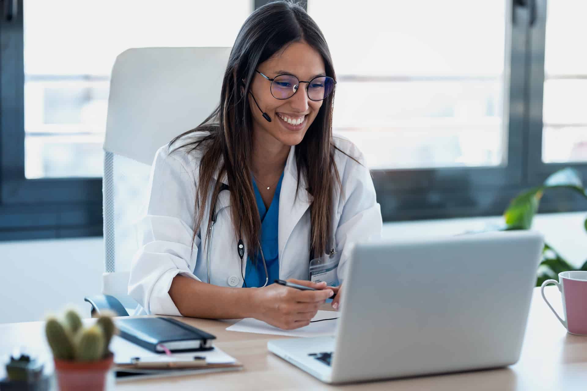 une jeune femme médecin installée à son bureau sourit en consultant son ordinateur