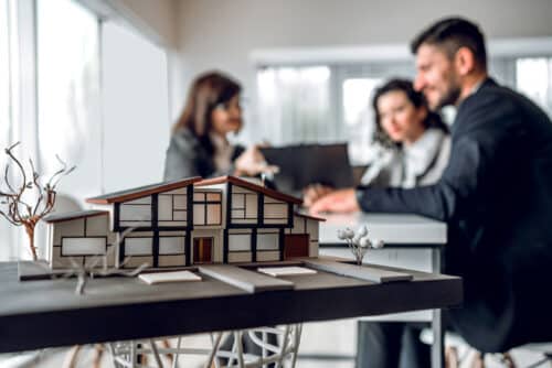 en avant plan une maquette de maison derrière un couple parle avec un architecte